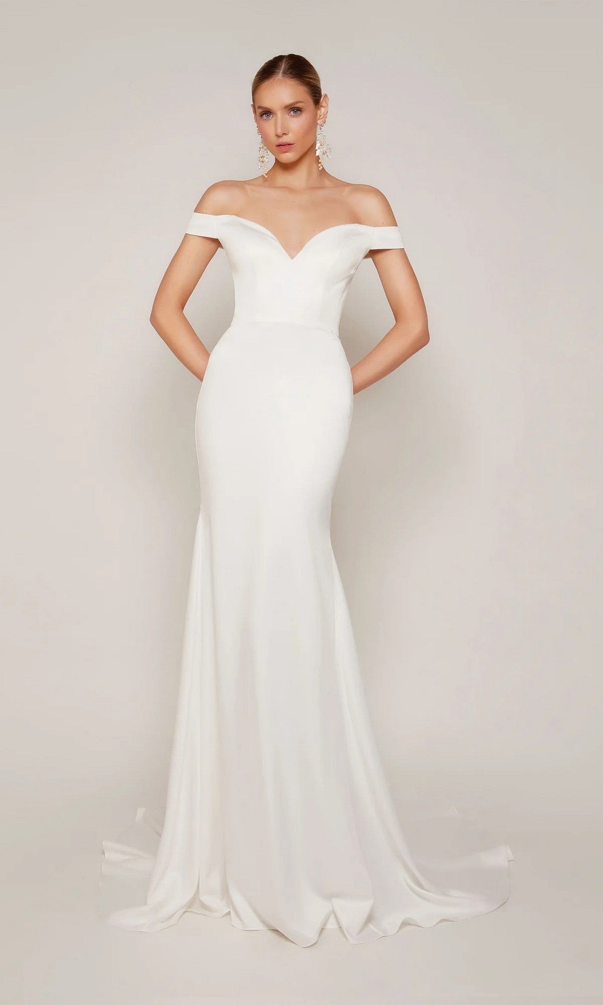 A woman stands against a plain background in the Alyce 7098—a long, fitted white formal dress with off-the-shoulder sleeves and a slight train.
